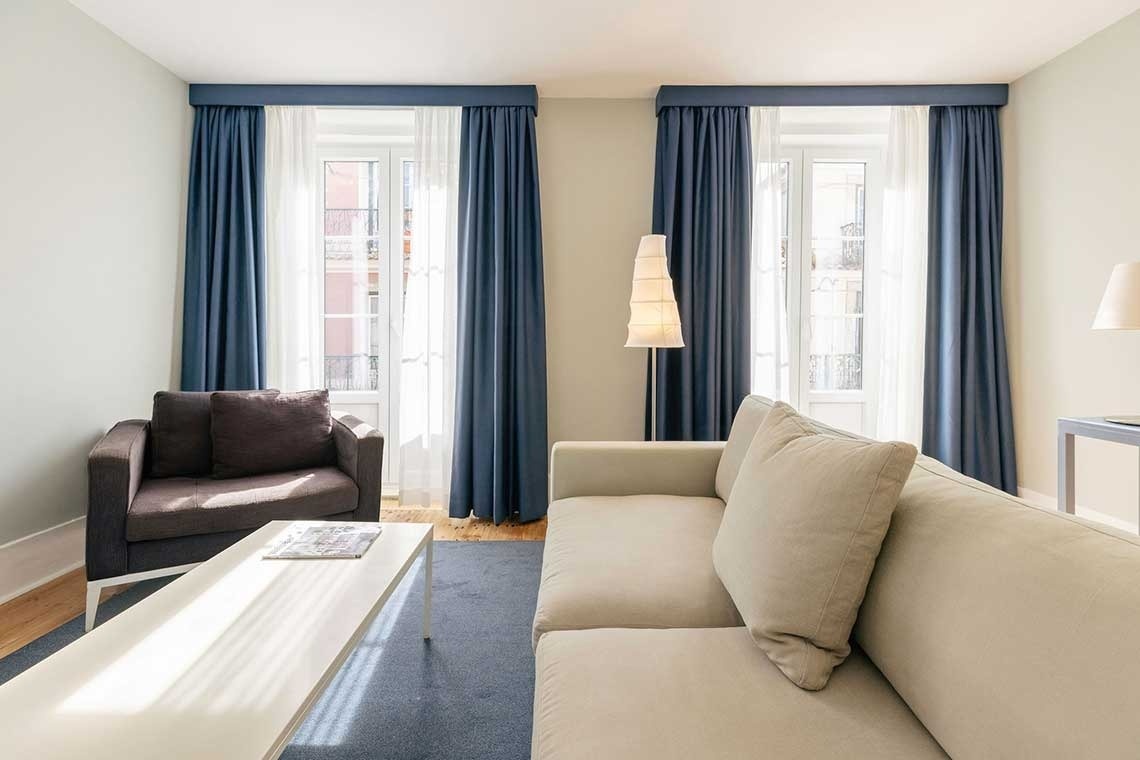 A bright living room features a cream-colored sofa, a dark armchair, a white coffee table, and two large windows framed by blue and sheer white curtains.
