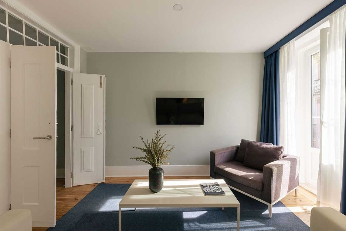 A bright and minimalist modern living room features light grey walls, wooden floors with a blue rug, a dark grey sofa, a white coffee table with a vase, and a wall-mounted TV, all illuminated by natural light streaming through a large window with blue and white curtains.