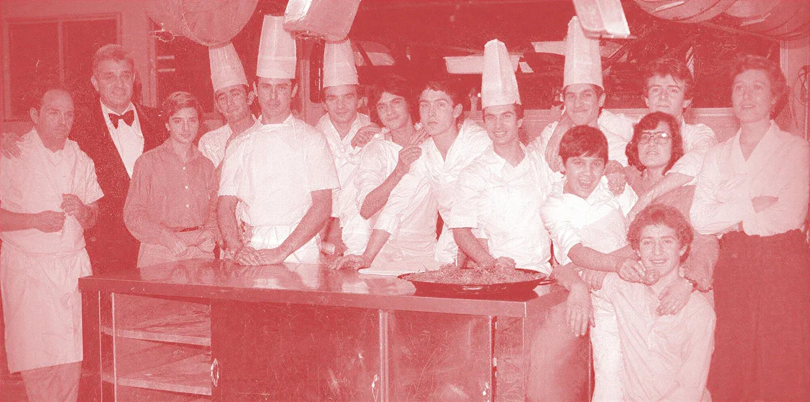 Un grupo de cocineros y estudiantes, la mayoría con gorros de chef, posa sonriente en una cocina alrededor de un gran plato de comida.