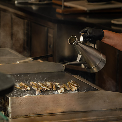 Chef adds oil to razor clams cooking on a griddle at a ESTIVAL: D Altafulla.