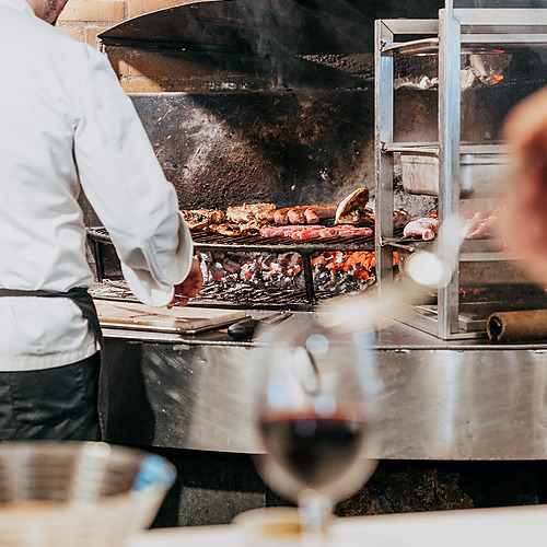 Chef grills ESTIVAL: D Altafulla meat and sausages over glowing coals in a restaurant.