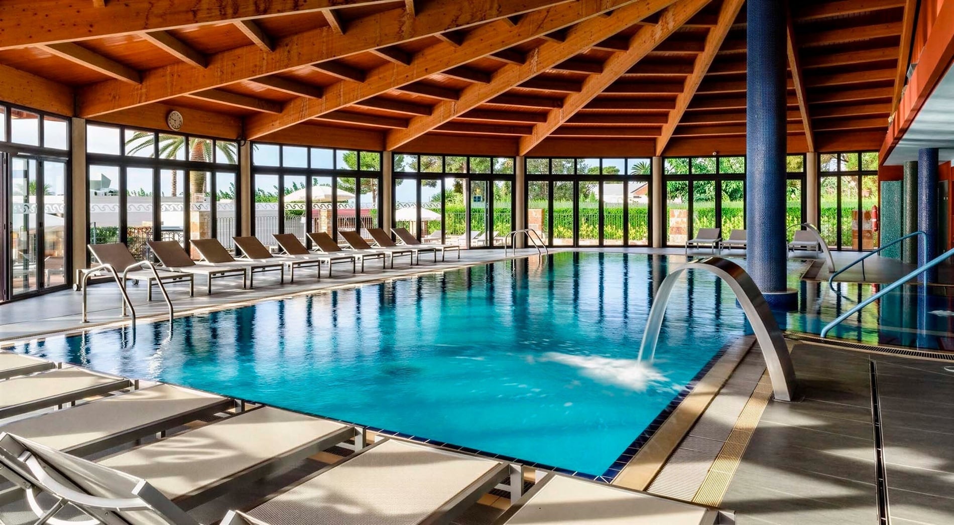 Una luminosa piscina cubierta cuenta con un impresionante techo de vigas de madera, grandes ventanales con vistas al exterior y una cascada de agua decorativa.