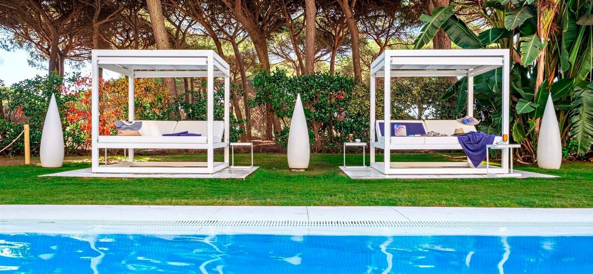 Two white outdoor daybeds with privacy screens are situated on a green lawn by a swimming pool, backed by a lush forest and tropical foliage.