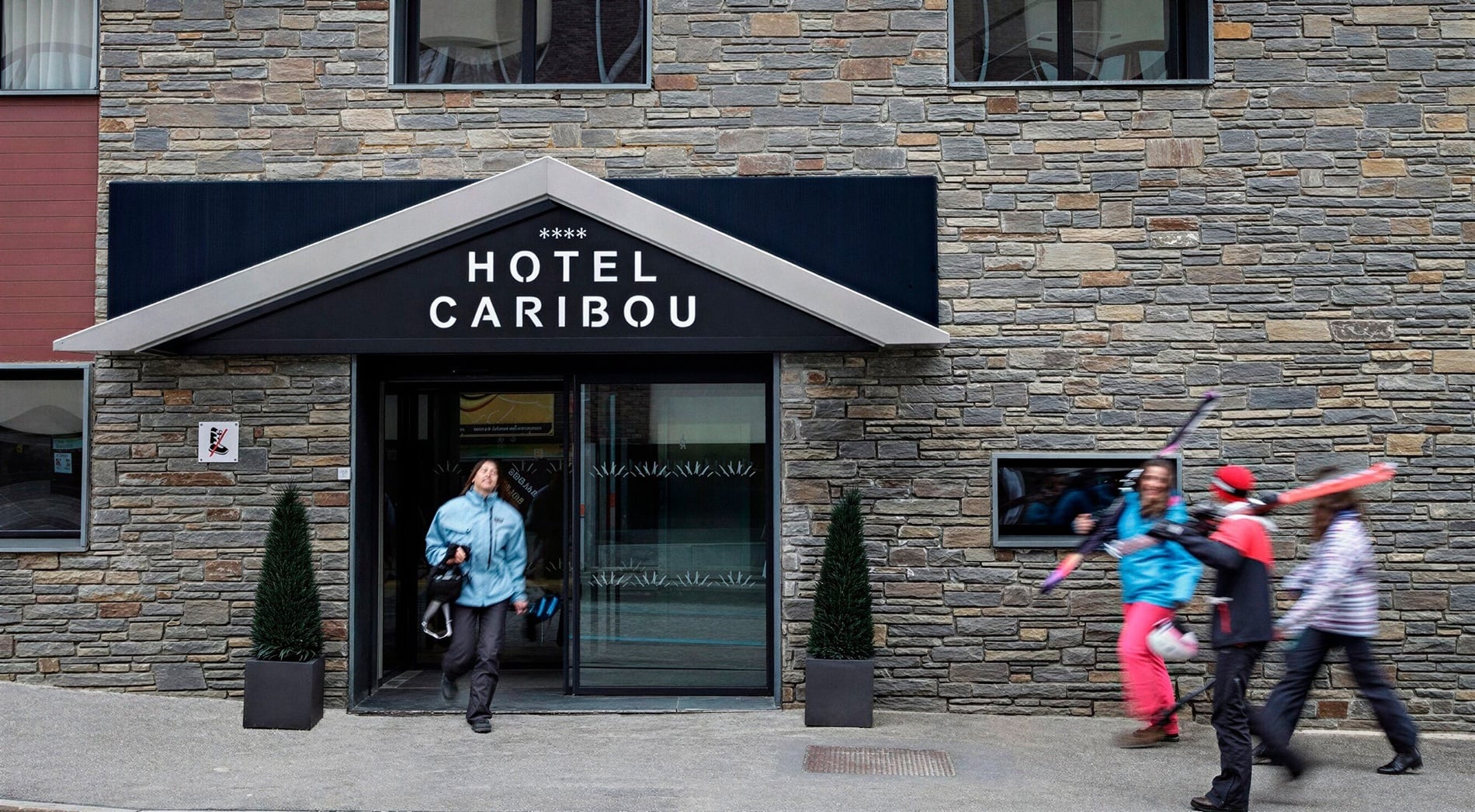 Blurred figures, some carrying skis, move past the stone facade entrance of the "Hotel Caribou."