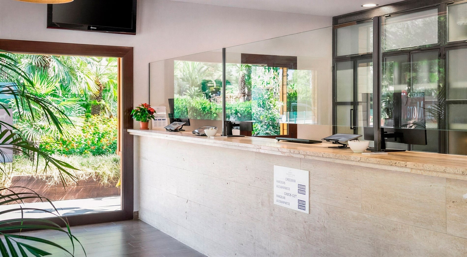 This image shows an outdoor restaurant patio with multiple tables and chairs set on a wooden deck, surrounded by trees, and offering a beautiful view of the clear blue sea under a bright sky.