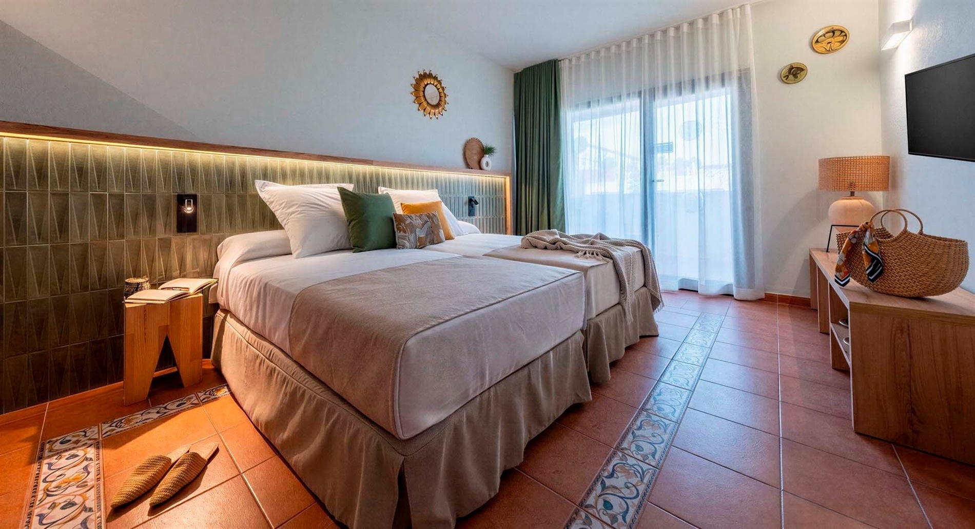 Una acogedora habitación de hotel con dos camas individuales, un cabecero iluminado y decoración en tonos tierra, complementada por un suelo de baldosas de terracota con acentos ornamentales.