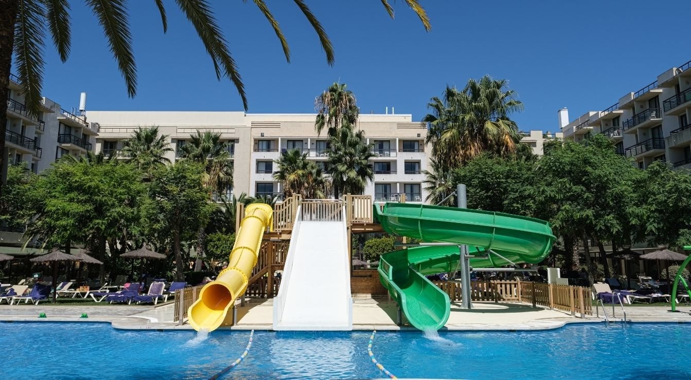 Eine Hotelanlage mit einem Poolbereich mit Wasserrutschen, umgeben von Palmen und Bäumen unter strahlend blauem Himmel.