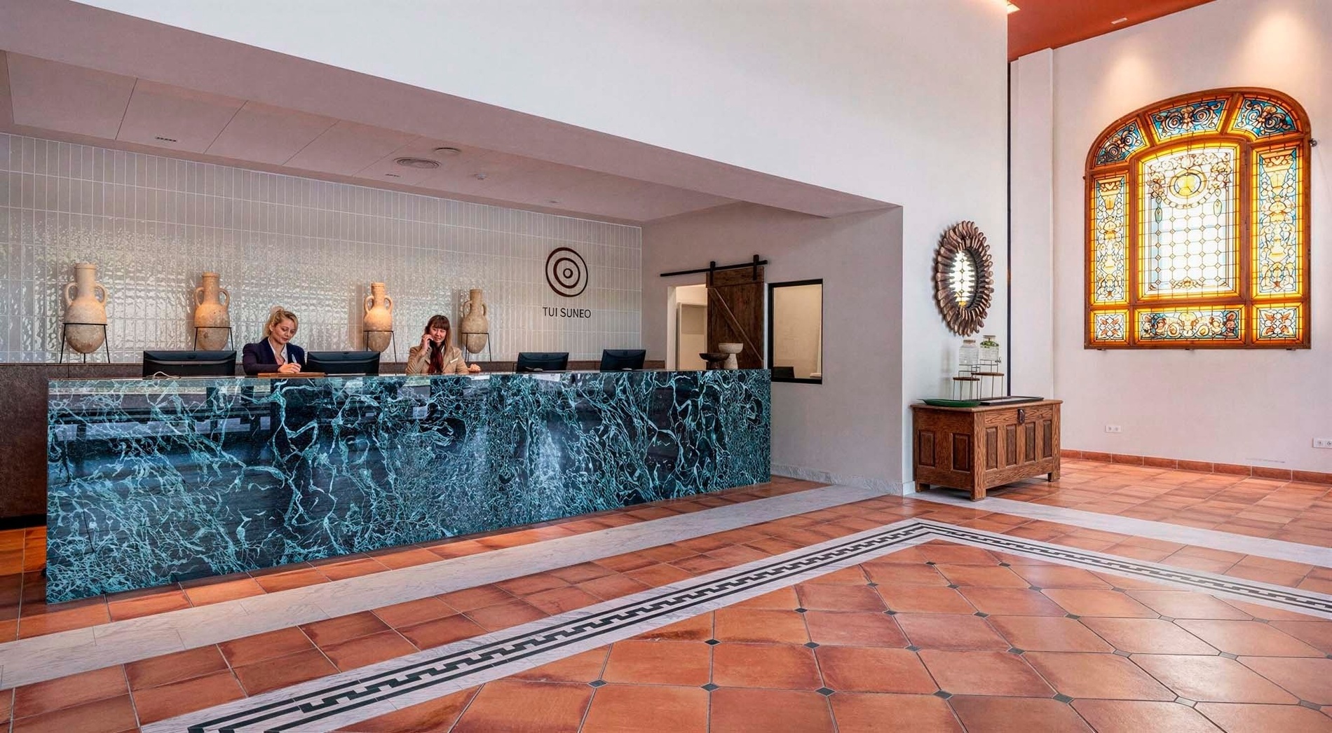 Two receptionists work behind a green marble desk in a hotel lobby featuring terracotta floor tiles, decorative urns, and a large stained-glass window.