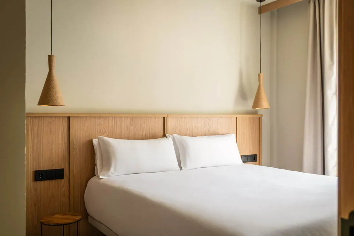 Habitació d'hotel moderna amb llit doble, capçal de fusta i llums penjants. ESTIVAL: Corporativa.