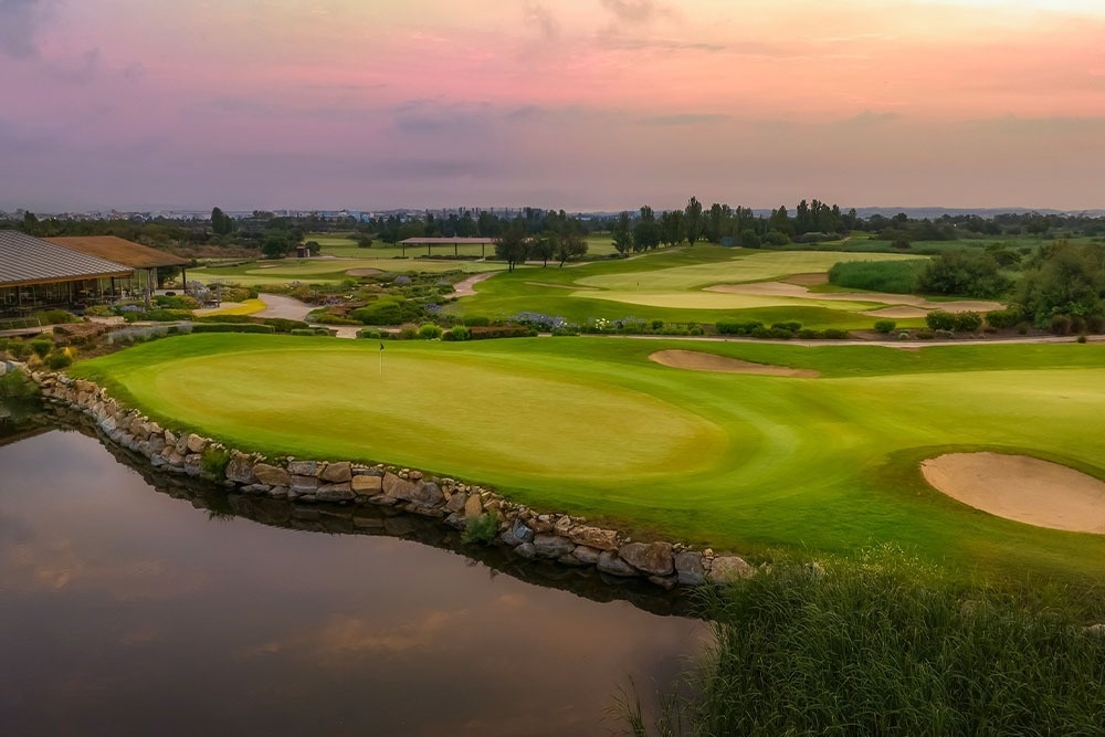 ca: Vista panoràmica d'un camp de golf amb un estany en primer pla i un cel amb colors de capvespre.