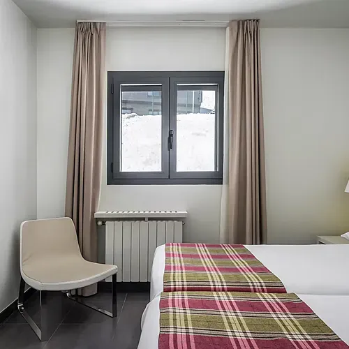 Cozy hotel room with twin beds, a TV, a chair, and a window with snowy views. Hotel Cristina | Andorra | Estival Group | Official Web