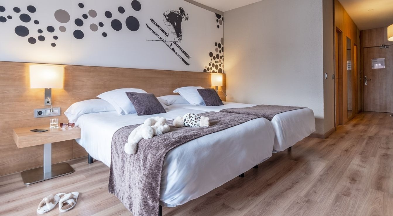 A modern hotel room features two neatly made single beds with dark accent pillows and throws, a light wood headboard, a decorative wall graphic of circles and a skier, and a light wood floor, with a bedside table holding a phone, glass of water, and sunglasses.