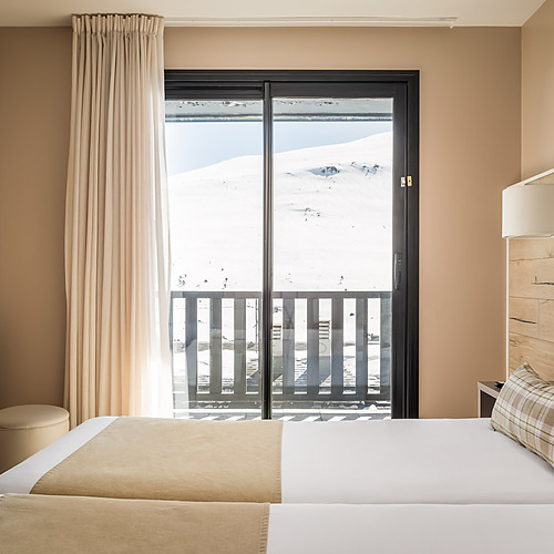 Hotel room with twin beds, balcony view of snowy mountains, and light beige walls. ESTIVAL: Corporativa