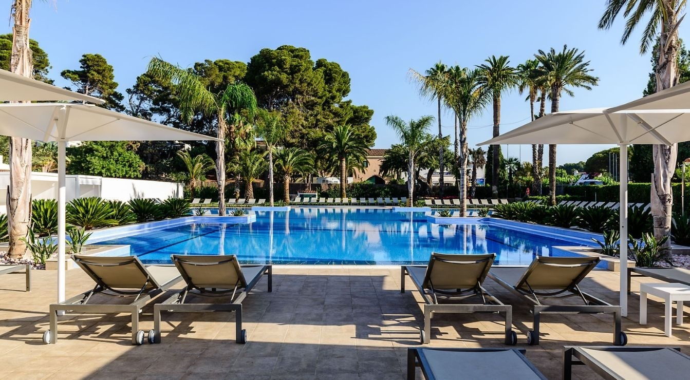 A large, rectangular swimming pool with clear blue water is surrounded by lounge chairs, white umbrellas, and abundant palm trees and greenery under a bright, clear sky.