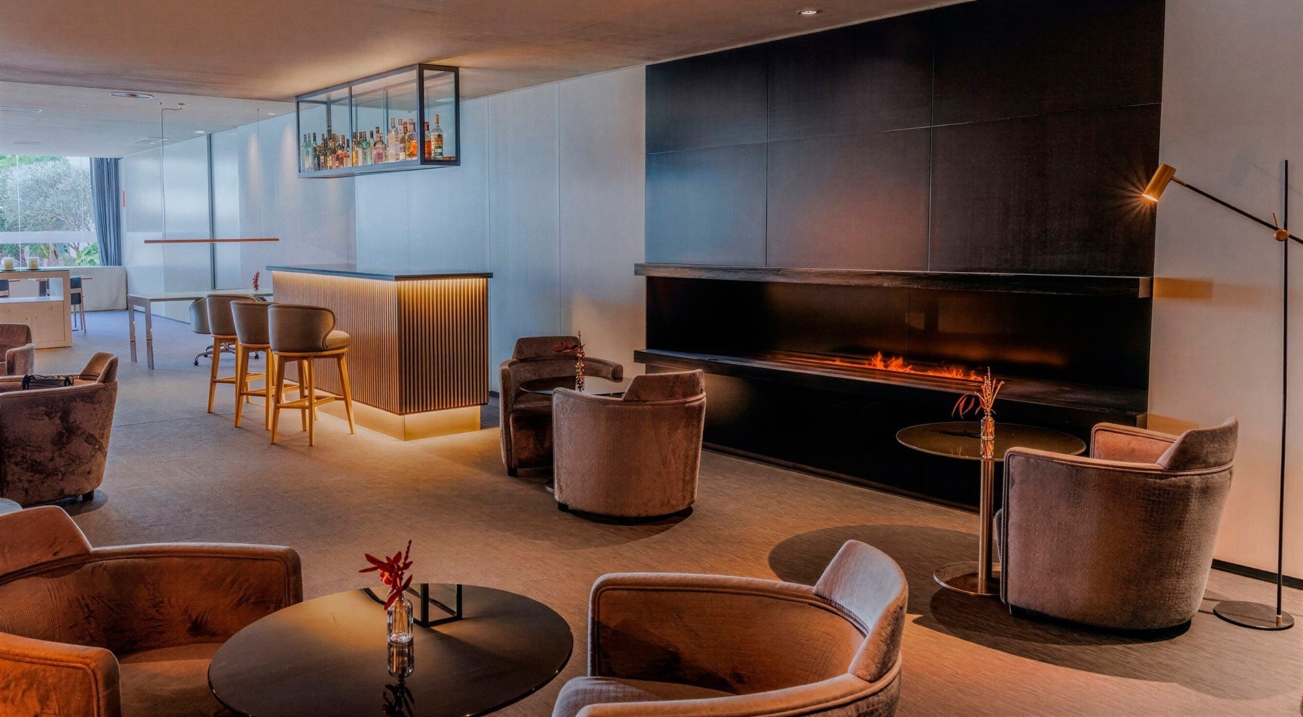 This image shows a sophisticated lounge area featuring a modern bar with backlit slatted wood, a long, built-in fireplace, and several plush armchairs arranged around small tables.