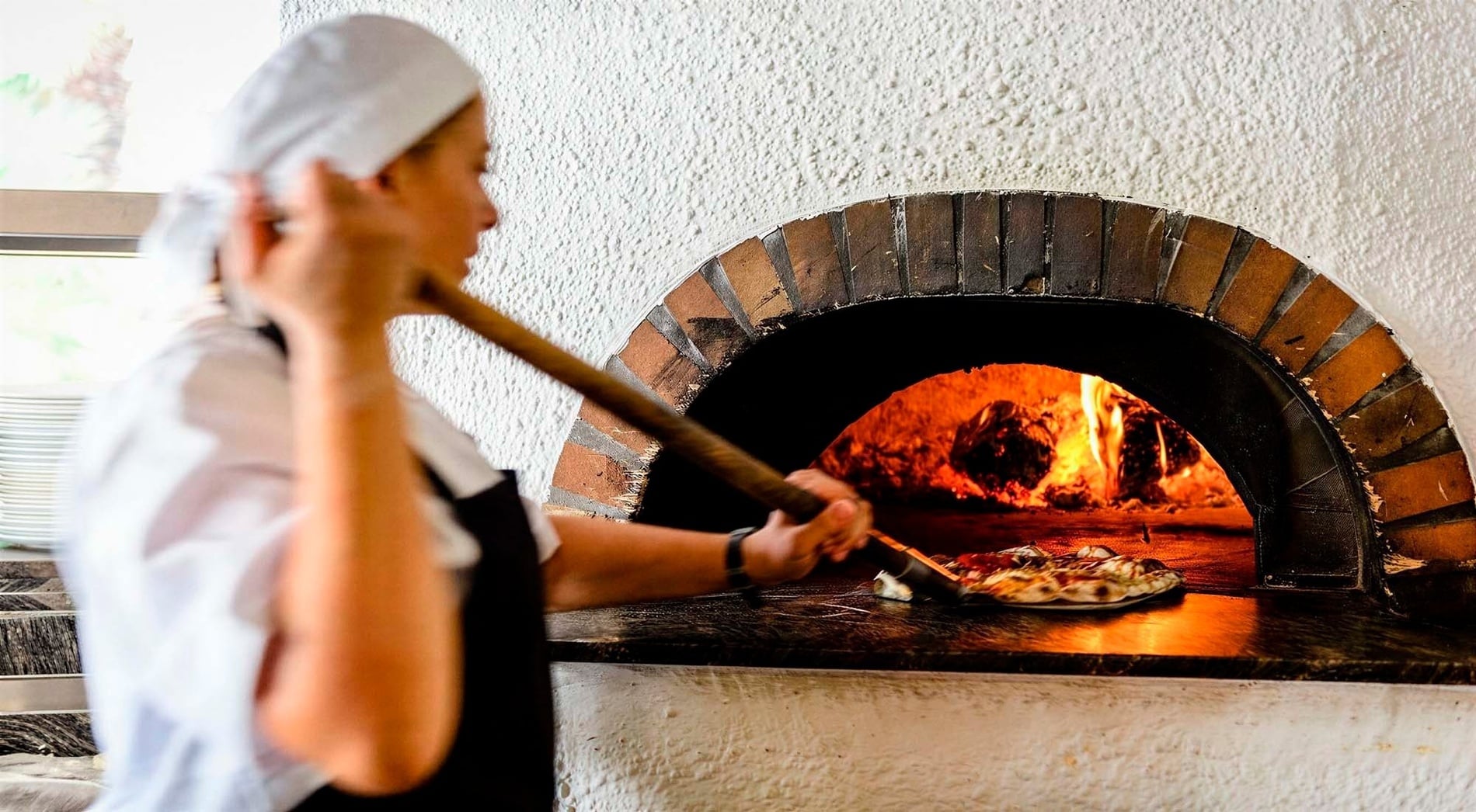Une personne enfourne une pizza dans un four à bois à l'aide d'une pelle.