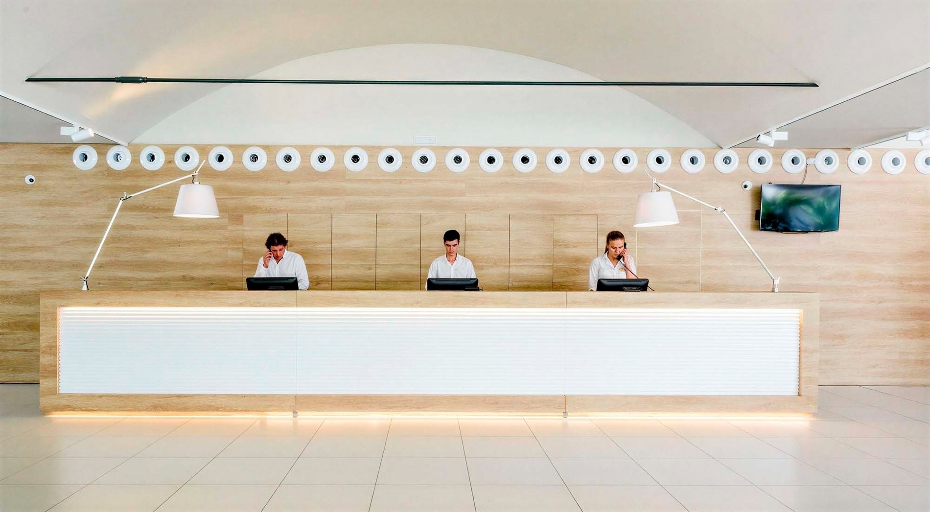 En una moderna recepción de hotel, tres empleados atienden detrás de un mostrador blanco y de madera, con una hilera de relojes decorativos y dos lámparas de diseño en la pared.