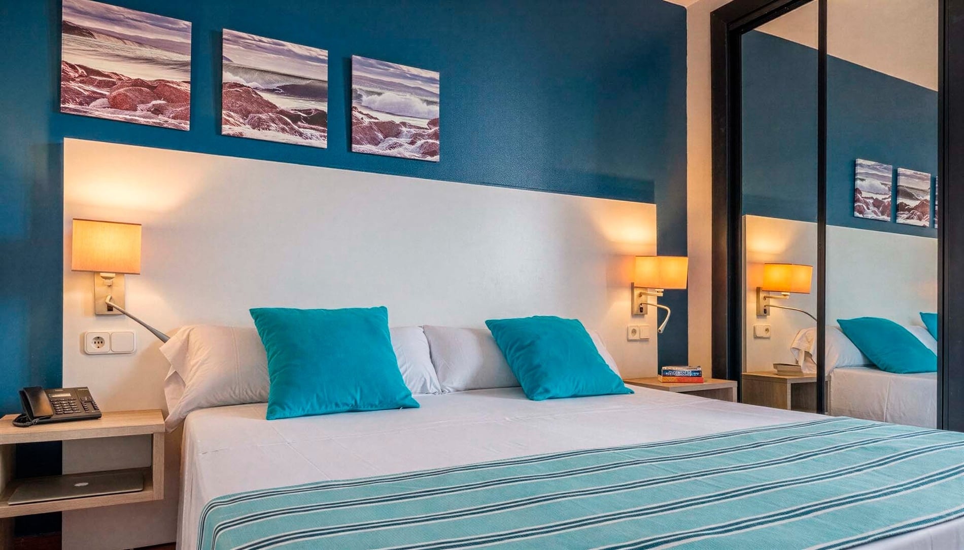 A modern bedroom features a large bed with white linens and teal accents, flanked by bedside tables with wall-mounted lamps, set against a two-tone wall with artwork and a mirrored wardrobe.