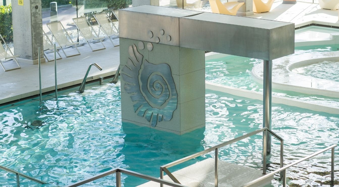 A modern indoor pool area is shown with clear blue water, a decorative pillar featuring a large shell design, and hot tub sections, under natural light.