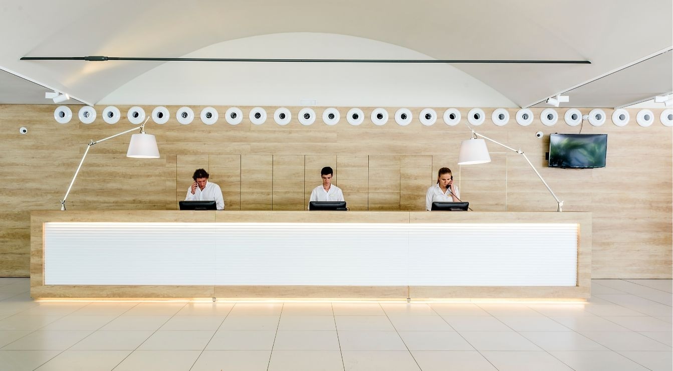 Three receptionists are busy attending to tasks behind a long, modern reception desk, under an array of circular decorative elements and stylish lamps.
