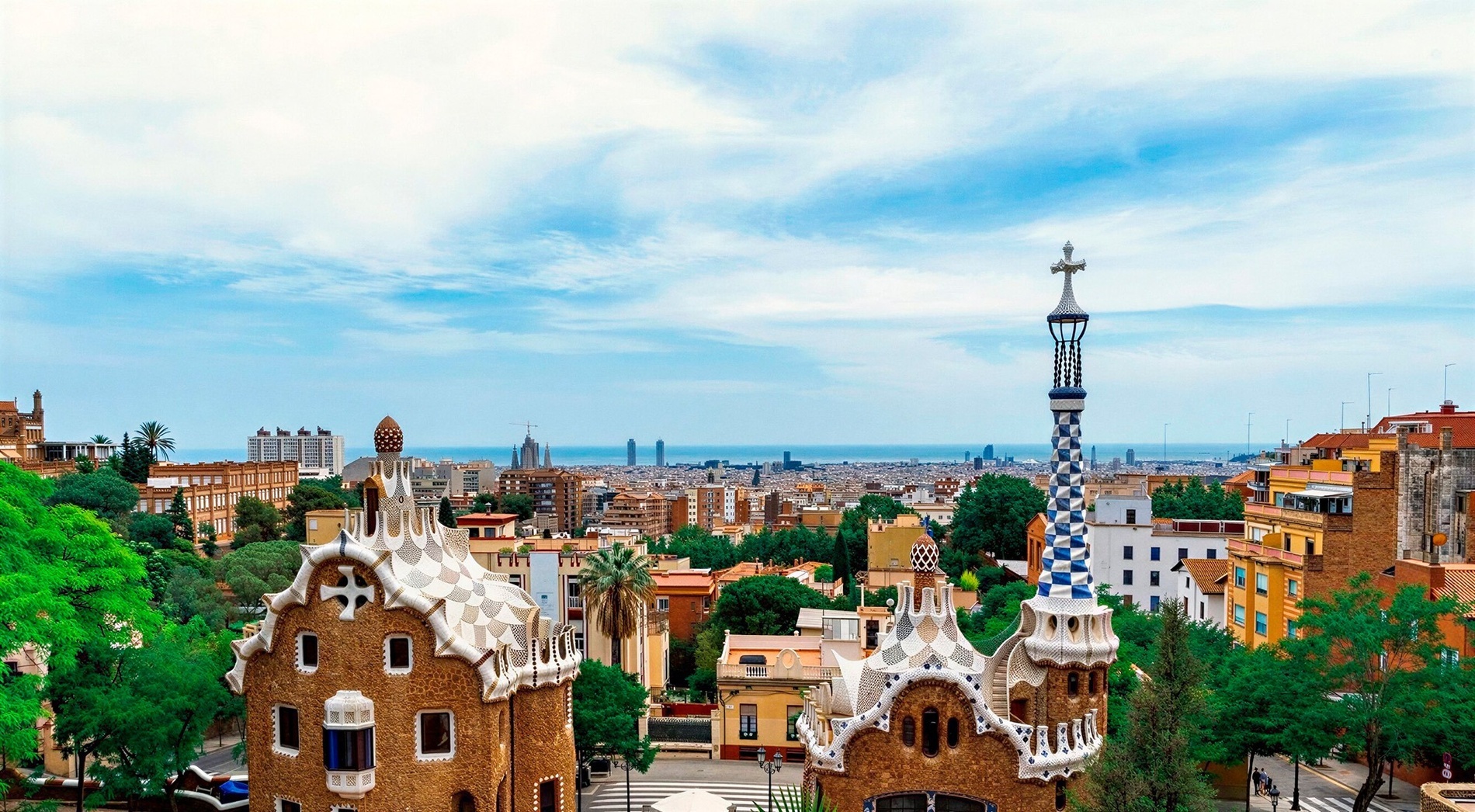 This image showcases the distinctive gingerbread-like buildings of Park Güell in Barcelona, overlooking a sprawling city with the Mediterranean Sea in the distance under a cloudy sky.