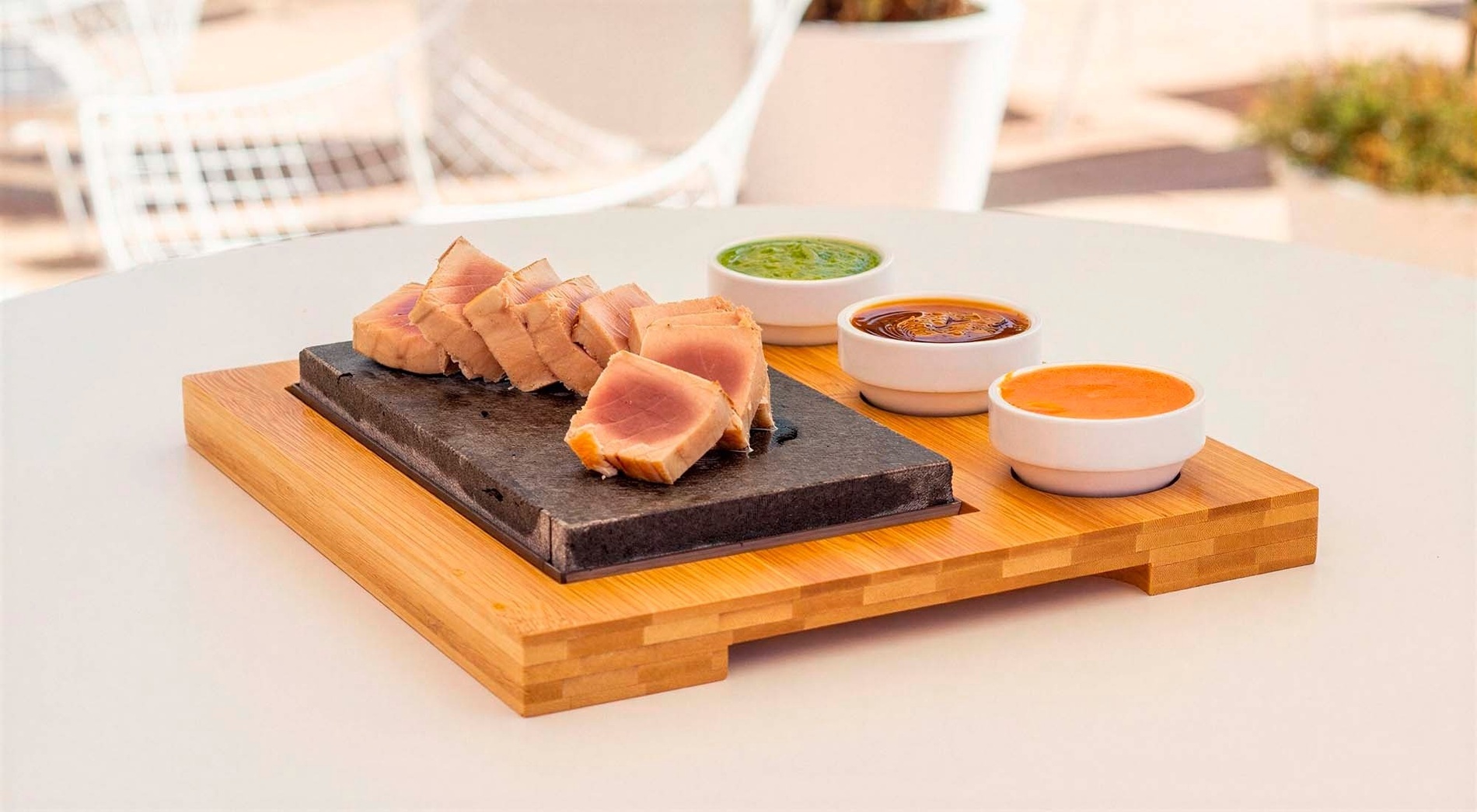 Tataki de atún sellado, dispuesto sobre una piedra volcánica en una bandeja de bambú, acompañado de tres salsas en cuencos individuales, sobre una mesa blanca.
