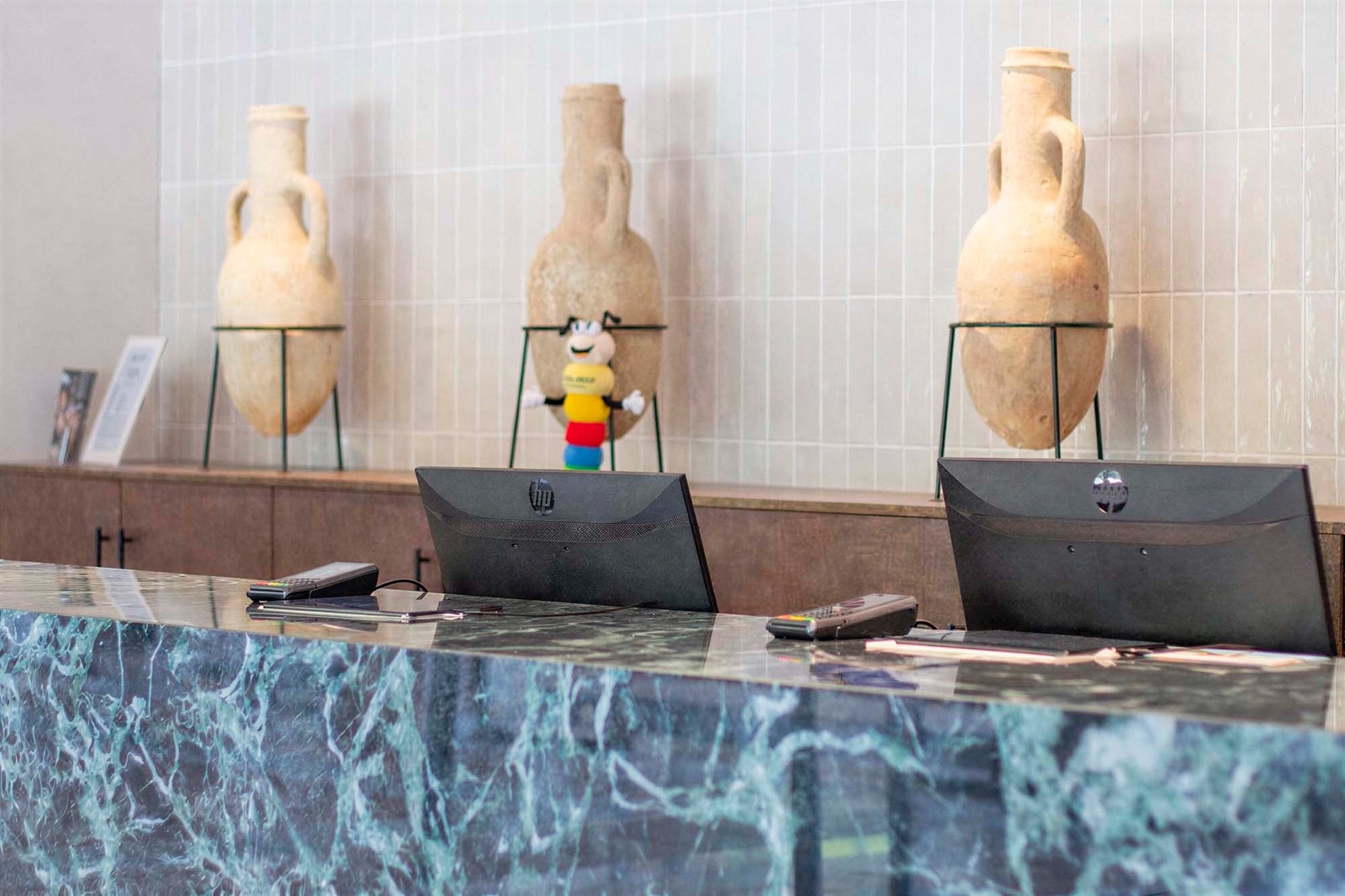 A woman in a blue polo shirt works at a reception desk with multiple computer monitors, set against a background featuring large decorative vases and a colorful toy.