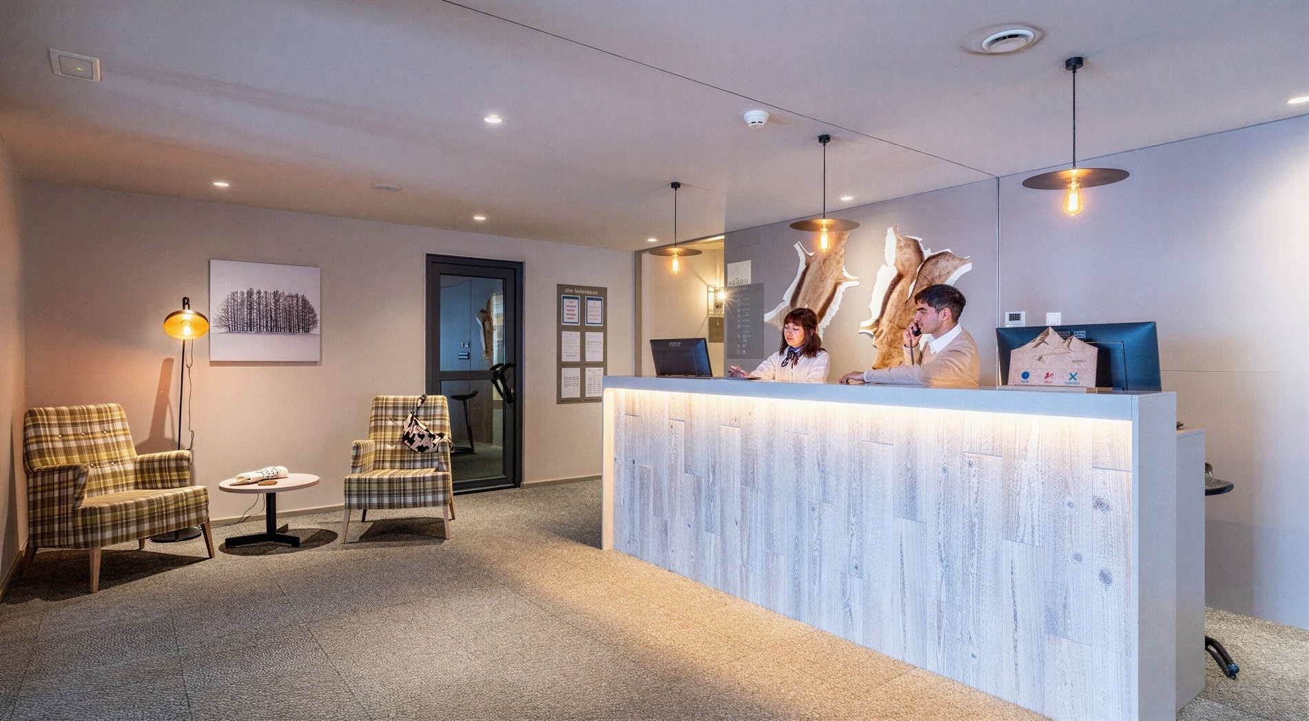 L'image présente une réception d'hôtel moderne avec deux employés derrière un comptoir éclairé et un petit coin salon meublé de fauteuils à carreaux.