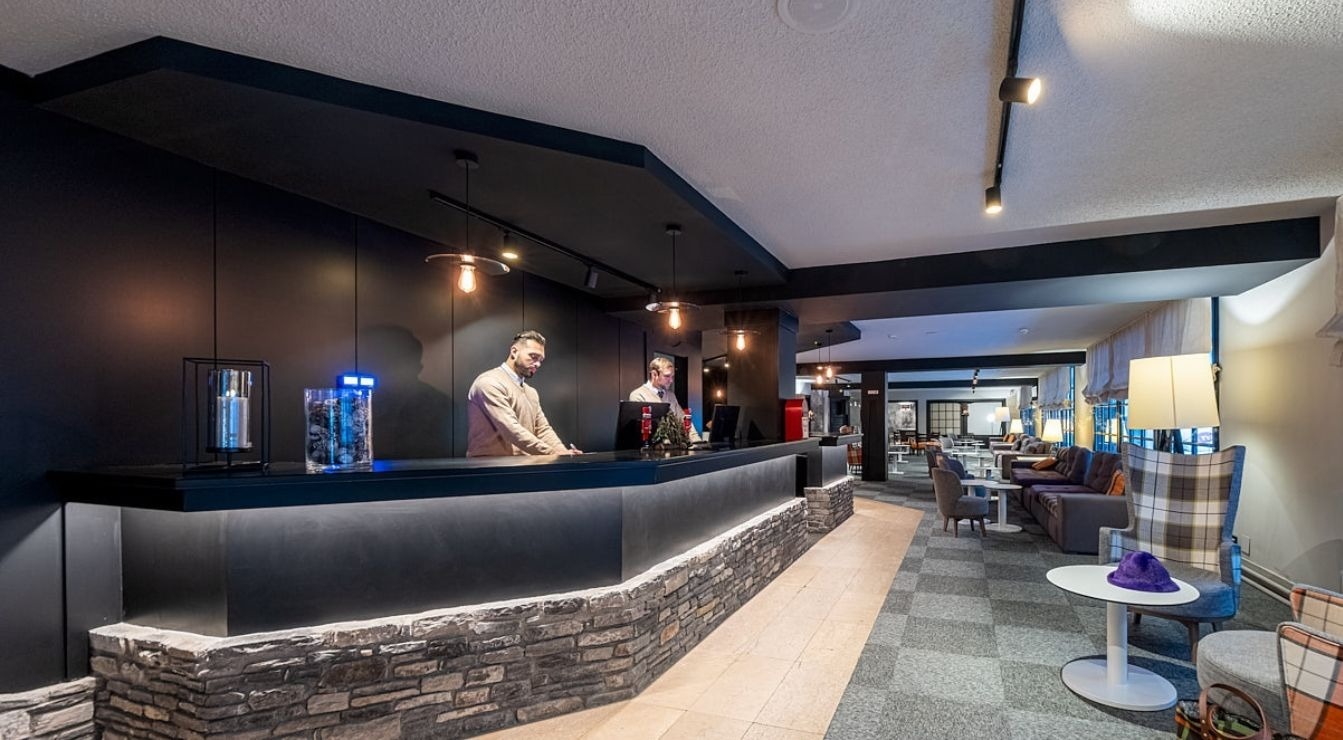 Two receptionists stand behind a stone and dark-paneled desk in a modern hotel lobby, with a comfortable lounge area visible to the right.