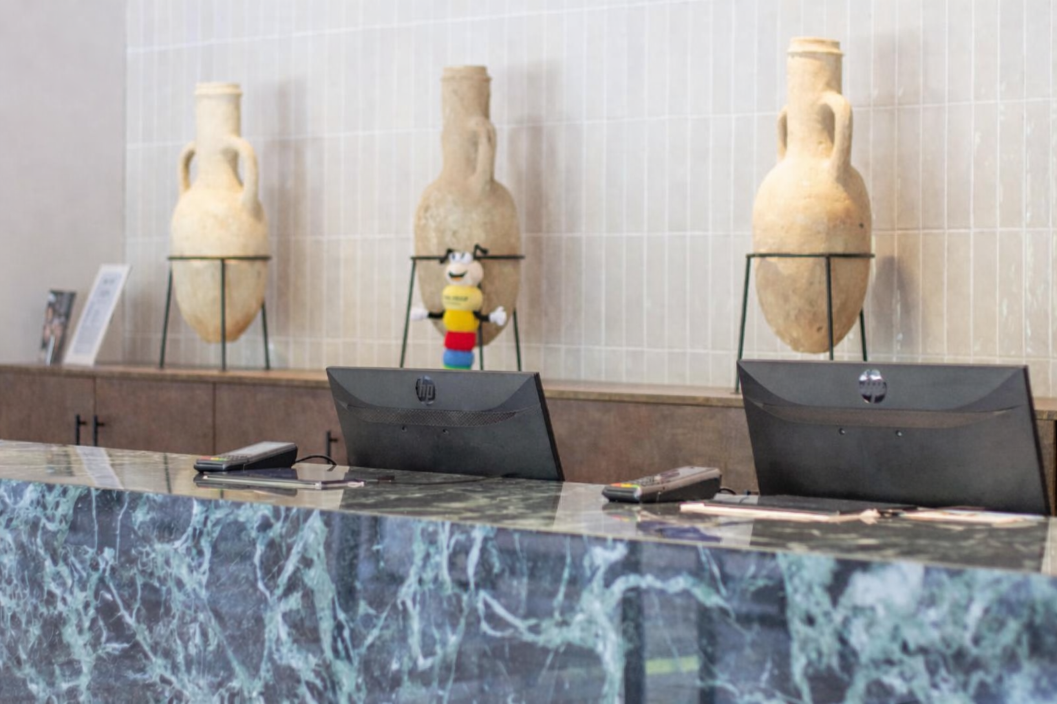 A woman in a blue polo shirt works at a reception desk with multiple computer monitors, set against a background featuring large decorative vases and a colorful toy.