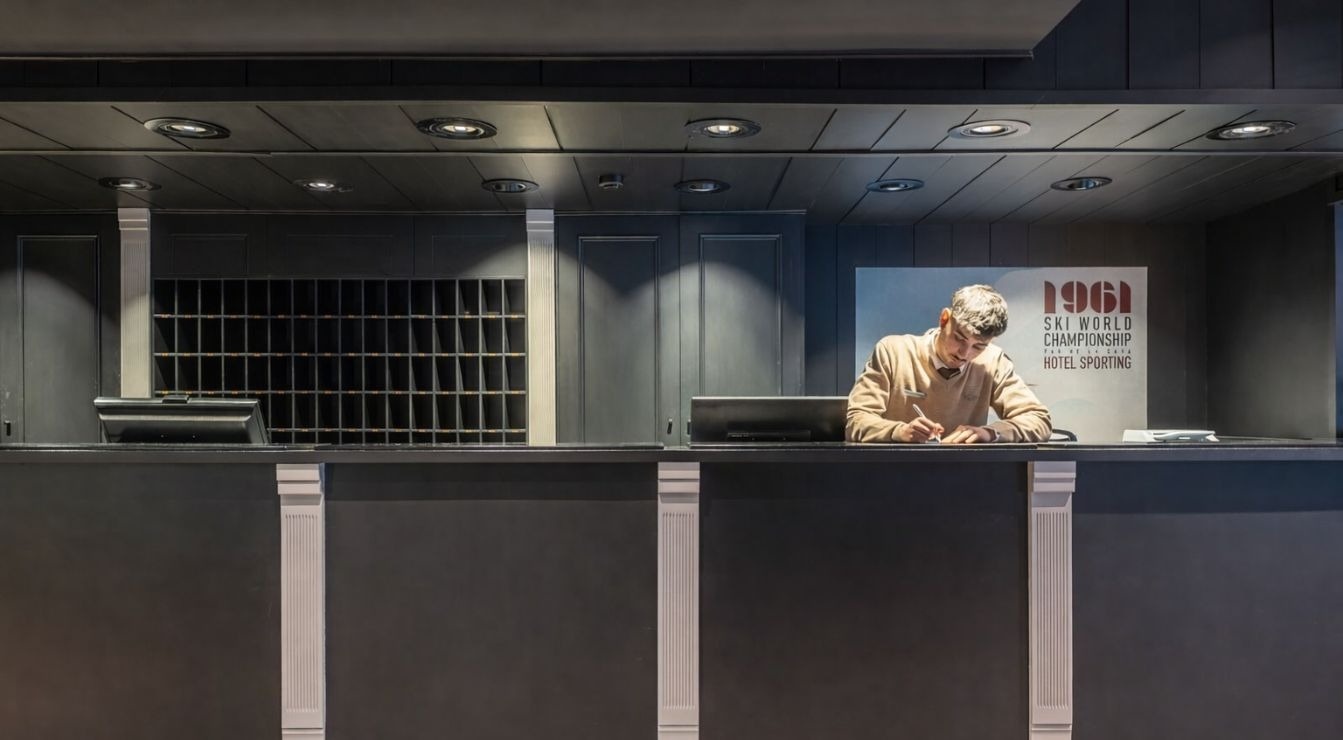 A man in a beige sweater is writing at a dark reception desk in a hotel lobby, which features a large pigeonhole mail rack and a sign referencing the "1961 SKI WORLD CHAMPIONSHIP HOTEL SPORTING."