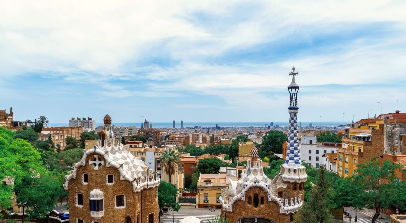 This image showcases the distinctive gingerbread-like buildings of Park Güell in Barcelona, overlooking a sprawling city with the Mediterranean Sea in the distance under a cloudy sky.