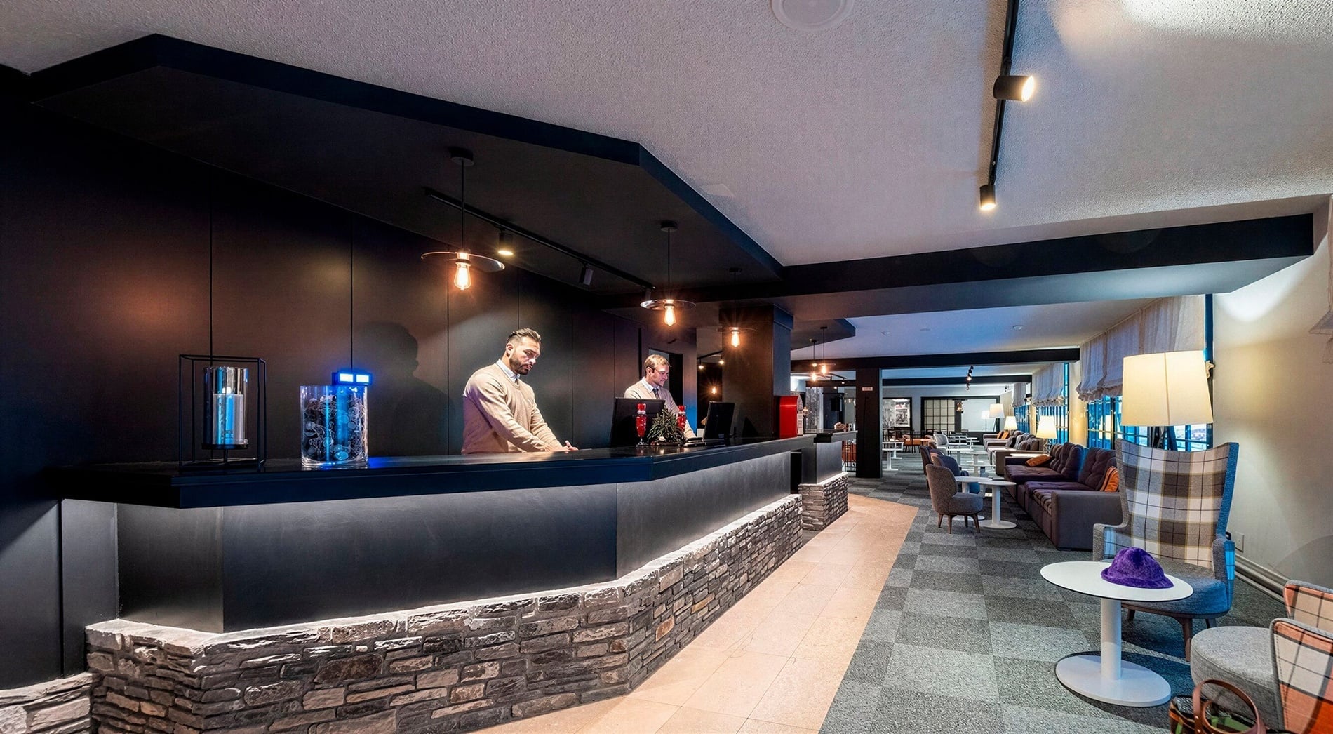 L'image présente un hall de réception d'hôtel élégant avec un comptoir en pierre illuminé, deux réceptionnistes et un salon confortable à droite.