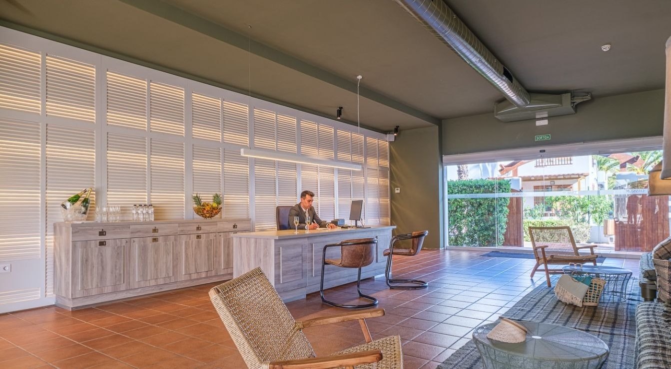 A modern reception area, possibly a hotel lobby, features a man working at a light-colored desk in front of an illuminated slatted wall, with terracotta tiled flooring and a glass door leading to an outdoor garden view.