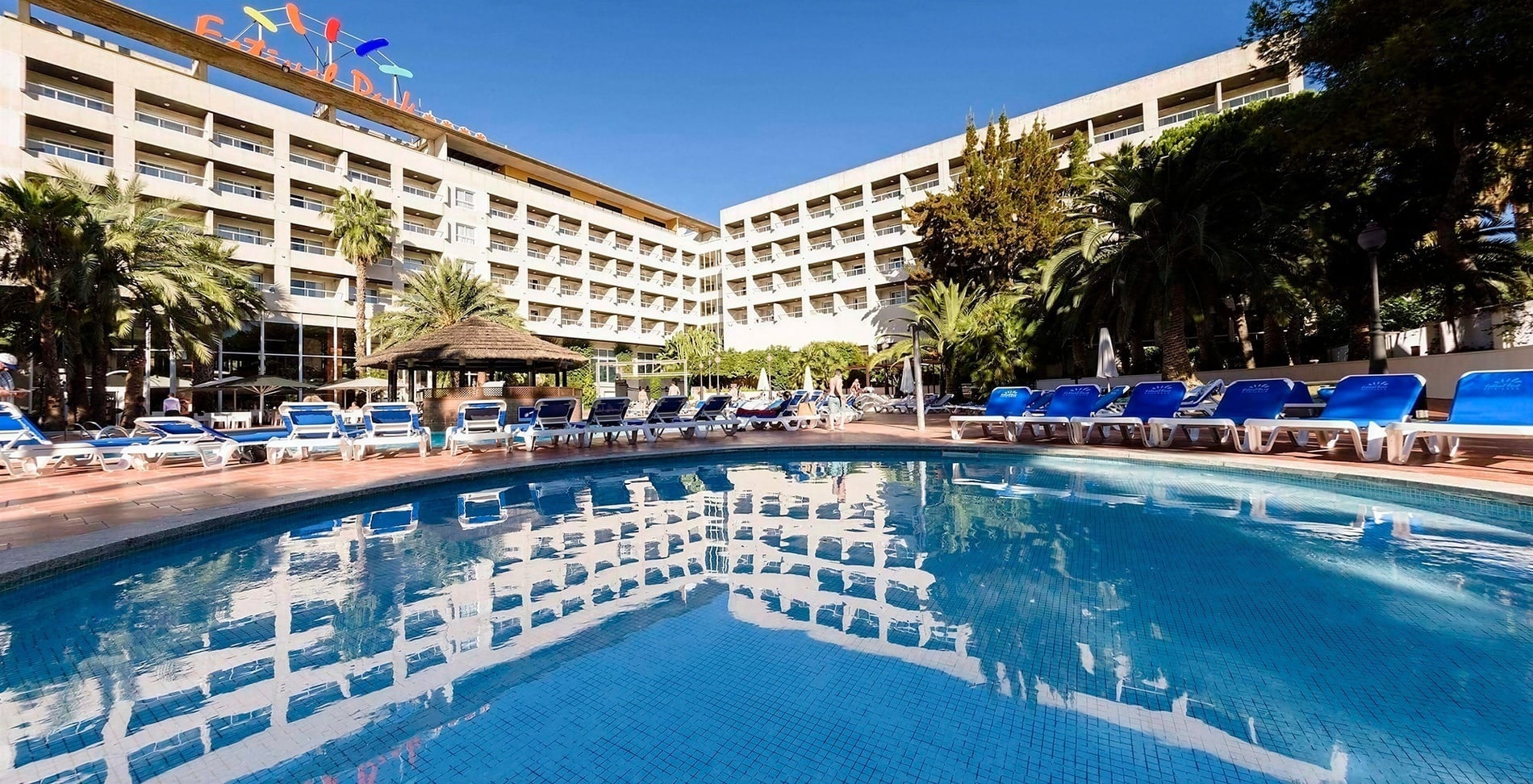 A large hotel with multiple balconies and colorful signage stands behind a sparkling blue swimming pool lined with sun loungers and surrounded by palm trees under a clear sky.