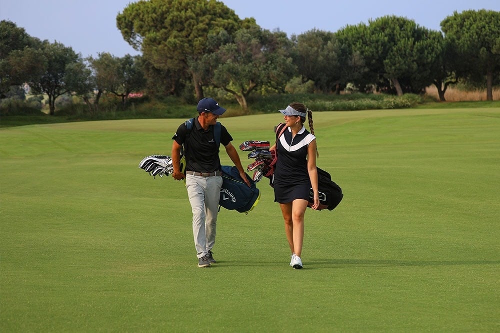 Ein Mann und eine Frau gehen mit ihren Golfausrüstungen über einen grünen Golfplatz, mit Bäumen im Hintergrund.