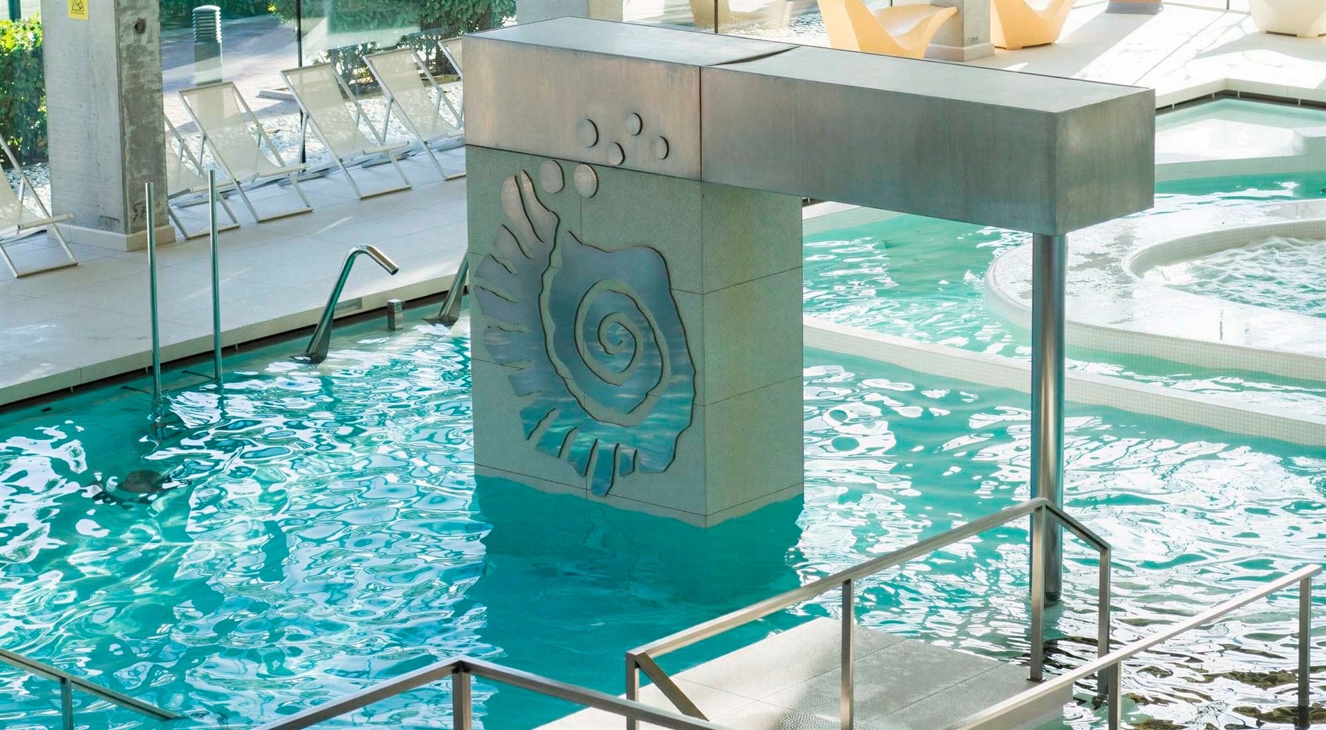 Ein lichtdurchfluteter Innenpool oder Spa-Bereich zeigt klares, türkisfarbenes Wasser, eine zentrale Säule mit Muschelornament und umgebende Liege- sowie Whirlpoolbereiche.