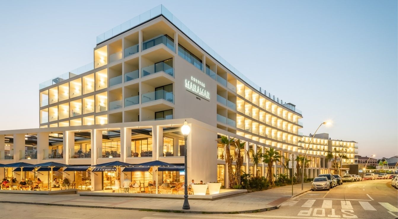 An aerial view captures a modern beachfront resort with multiple swimming pools, extensive sun loungers, and a street lined with parked cars, all set against the backdrop of a sandy beach and the blue ocean.