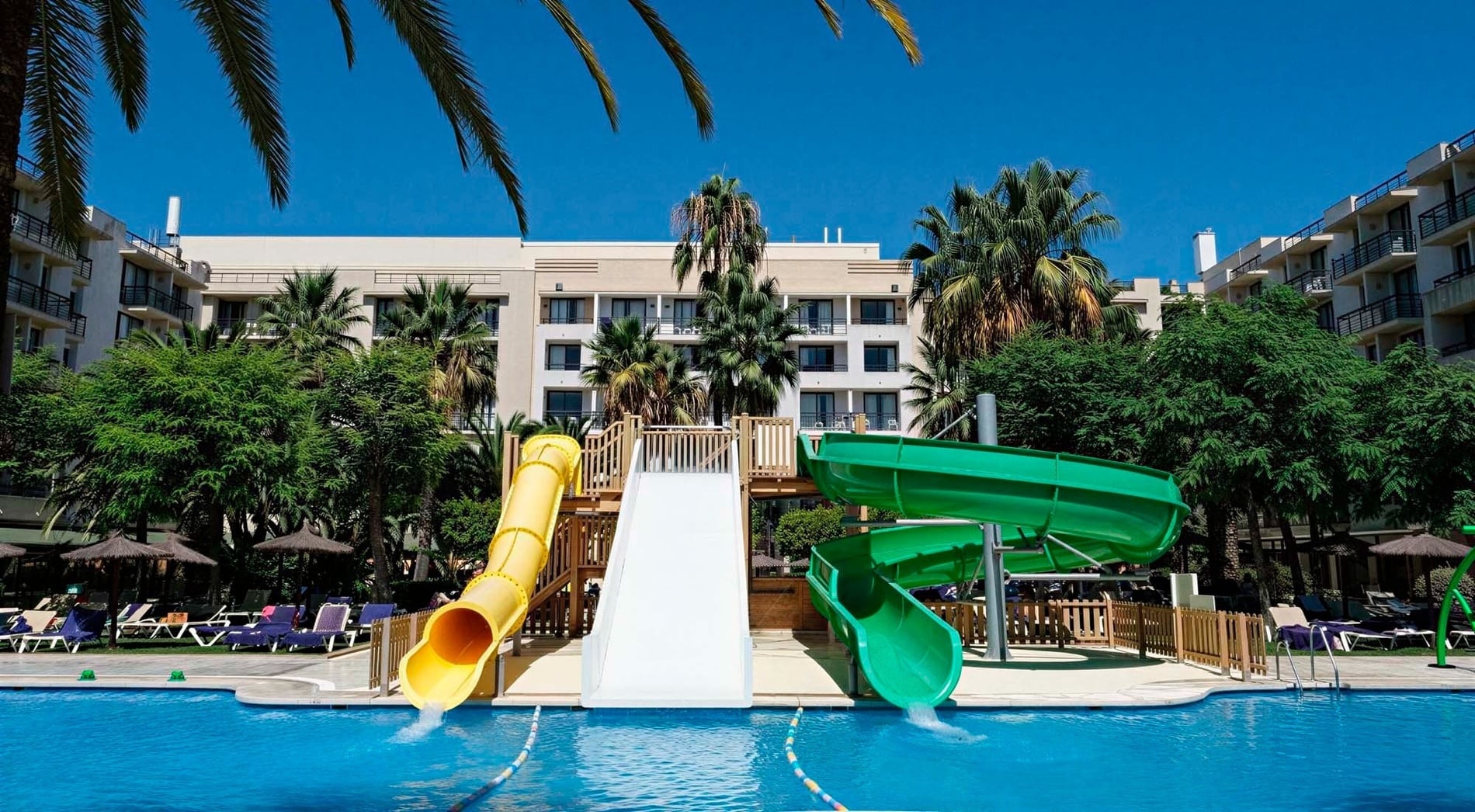 This vibrant image captures a resort swimming pool featuring yellow, white, and green water slides, set against a backdrop of a large hotel building and lush palm trees under a bright blue sky.