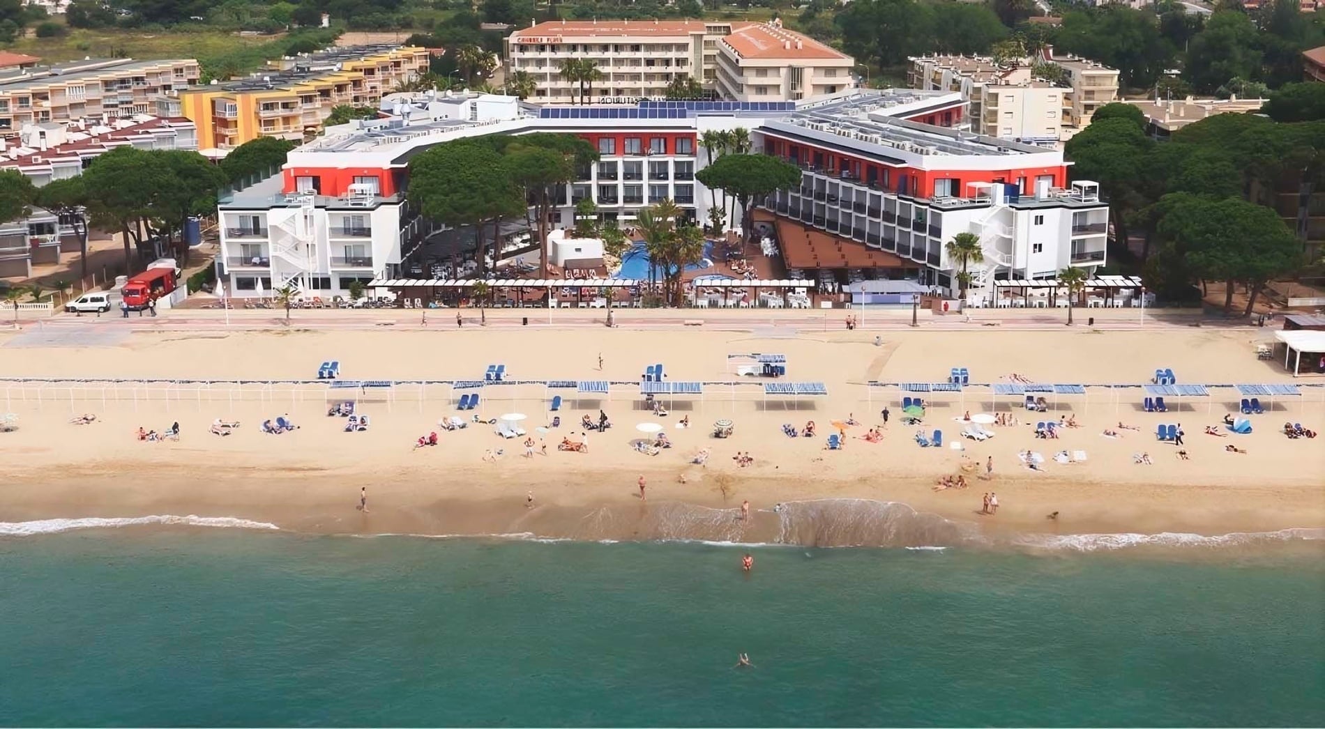 Eine Luftaufnahme zeigt einen belebten Strand mit Badegästen und Sonnenschirmen, direkt angrenzend an einen Hotelkomplex mit Pools und Palmen.