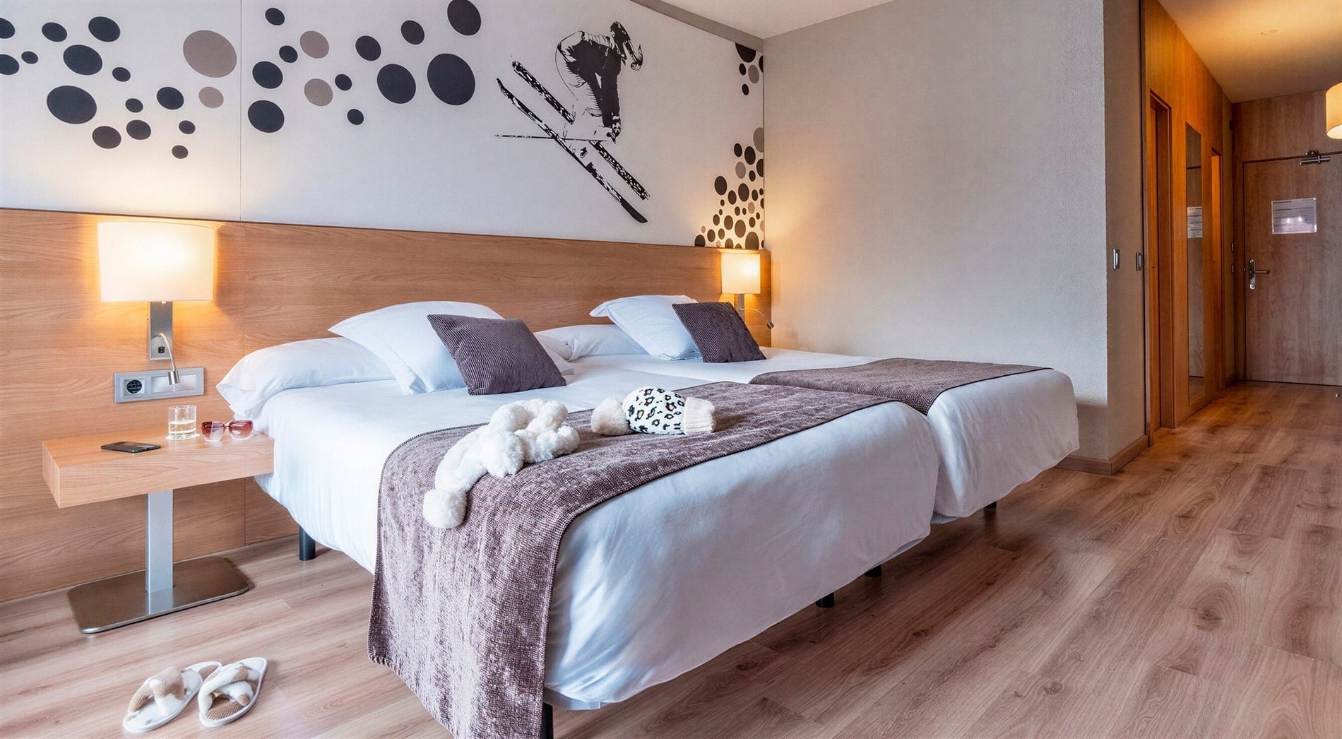 A modern hotel room features two neatly made single beds with dark accent pillows and throws, a light wood headboard, a decorative wall graphic of circles and a skier, and a light wood floor, with a bedside table holding a phone, glass of water, and sunglasses.
