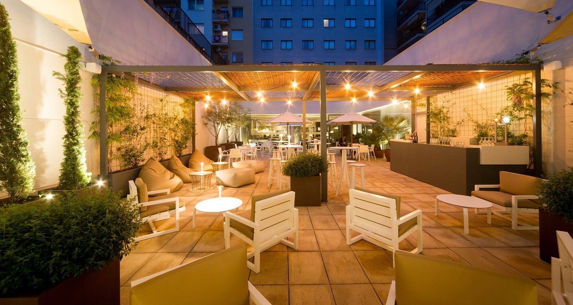 Two hotel staff members, a man and a a woman, are stationed behind a modern reception desk with a "HOTEL VILAMARÍ" sign, while a street scene with cars and a set of stairs are visible through large windows in the background.
