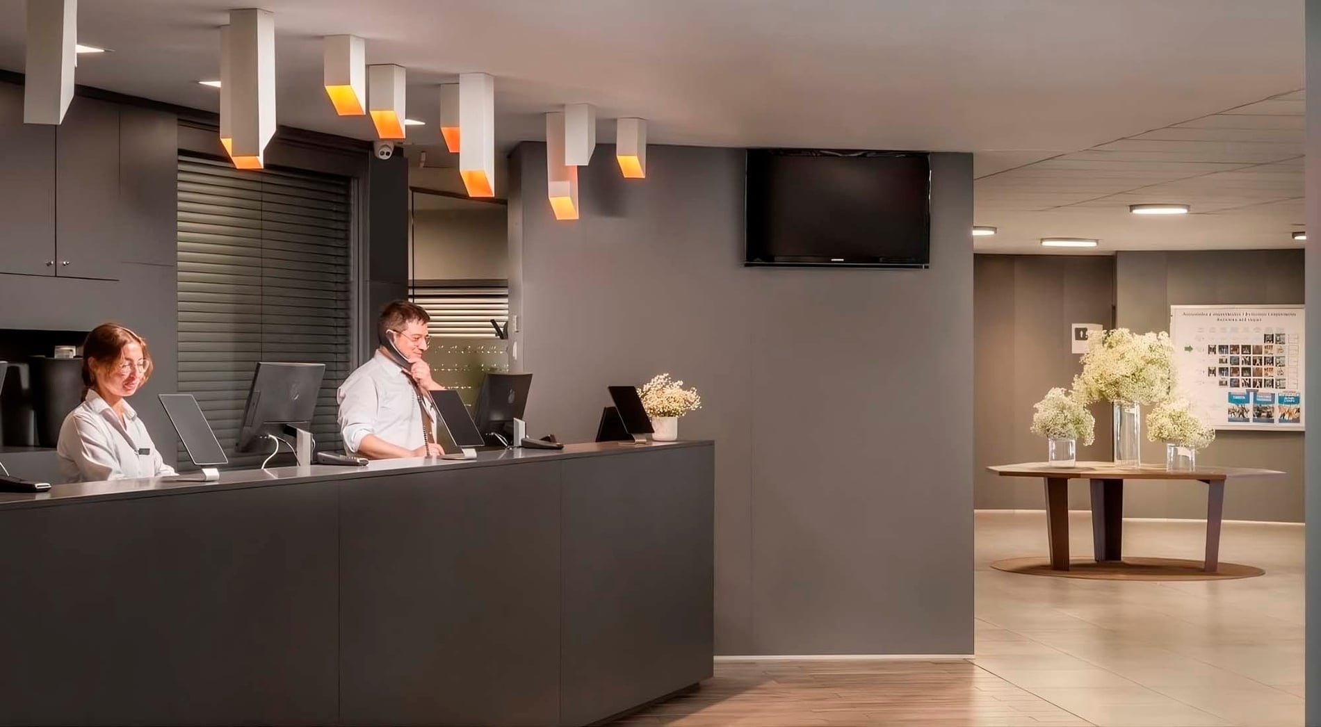 Two receptionists work at a modern dark grey desk under unique hanging lights, with a television mounted on the wall and a table adorned with white flowers in the background.
