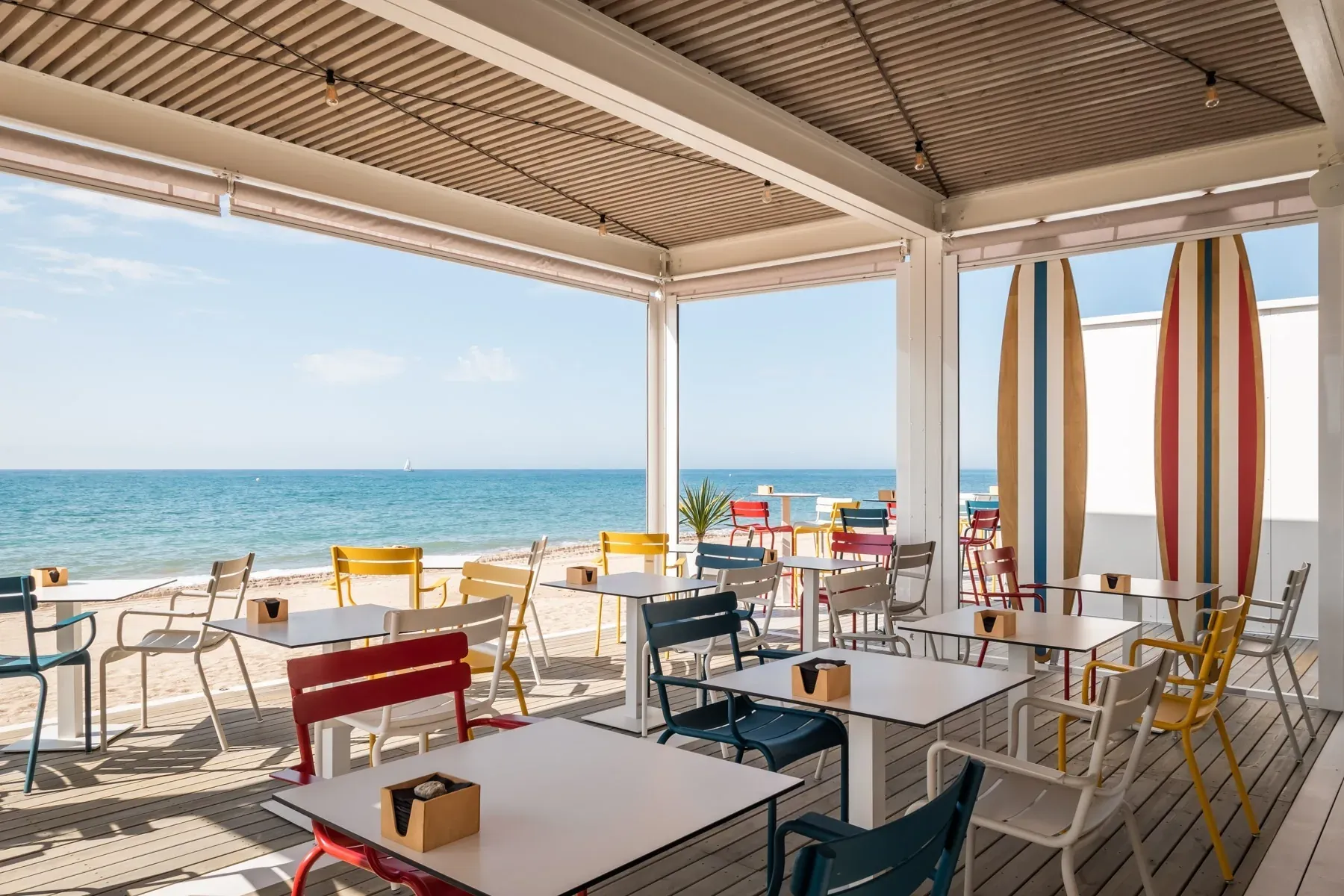 Strandrestaurant mit bunten Tischen & Stühlen, Meerblick, Surfbrettern. Ideal für ESTIVAL: Firmen.