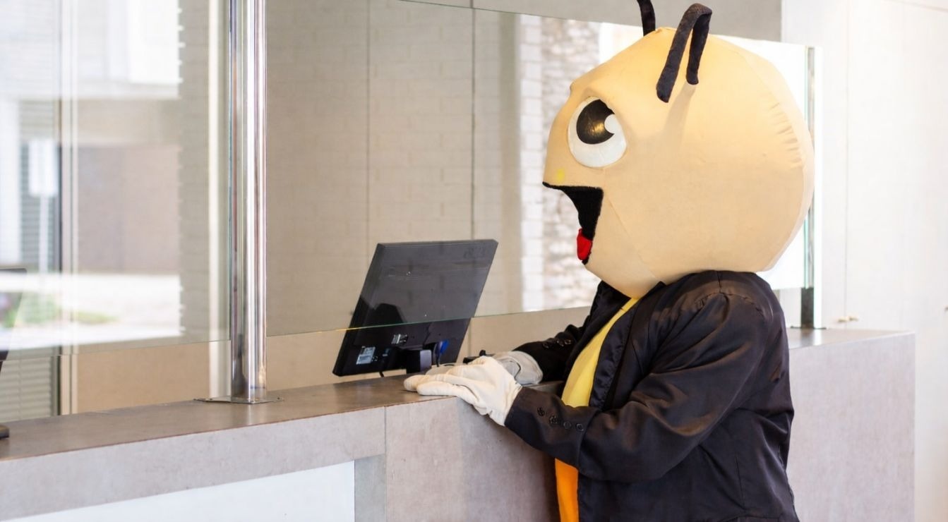 A person in an insect mascot costume, dressed in a suit and white gloves, stands at a counter and looks at a computer screen behind a glass partition.