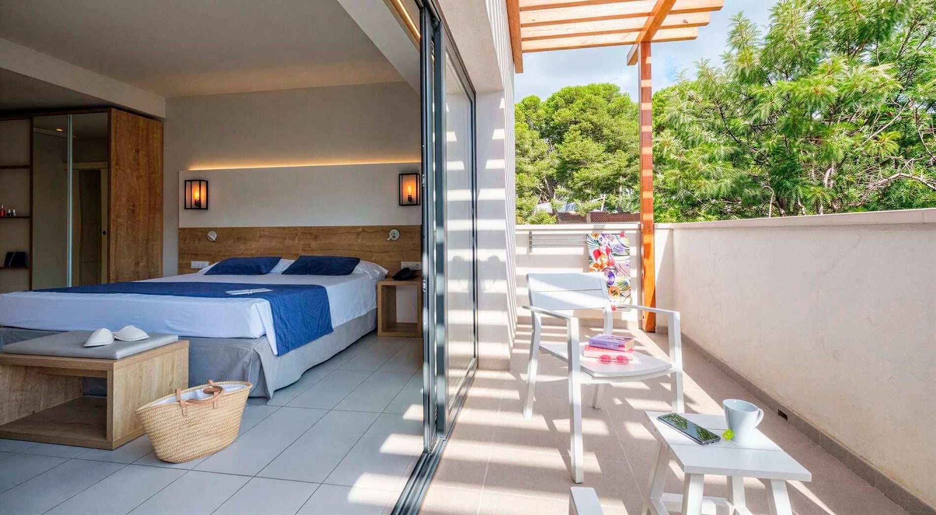 Ein helles, modernes Schlafzimmer mit einem Doppelbett und angrenzendem, sonnigen Balkon, der von einer Holzpergola beschattet wird und einen Blick ins Grüne bietet.