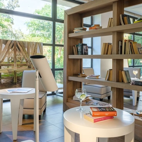 Un salon de lecture lumineux et moderne, avec de grandes baies vitrées donnant sur la verdure, est aménagé avec une bibliothèque en bois remplie de livres et des fauteuils confortables.