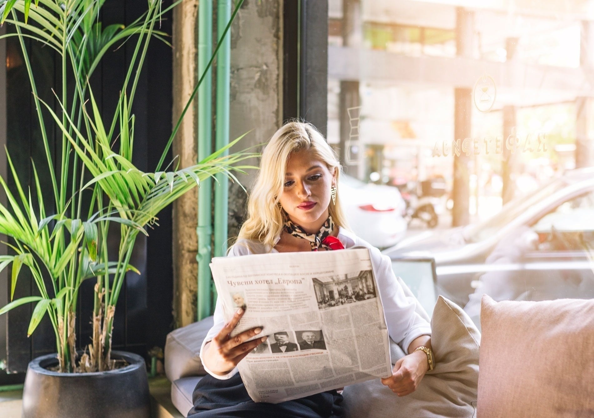 Una mujer rubia lee un periódico en un café luminoso, sentada junto a una planta y una ventana.
