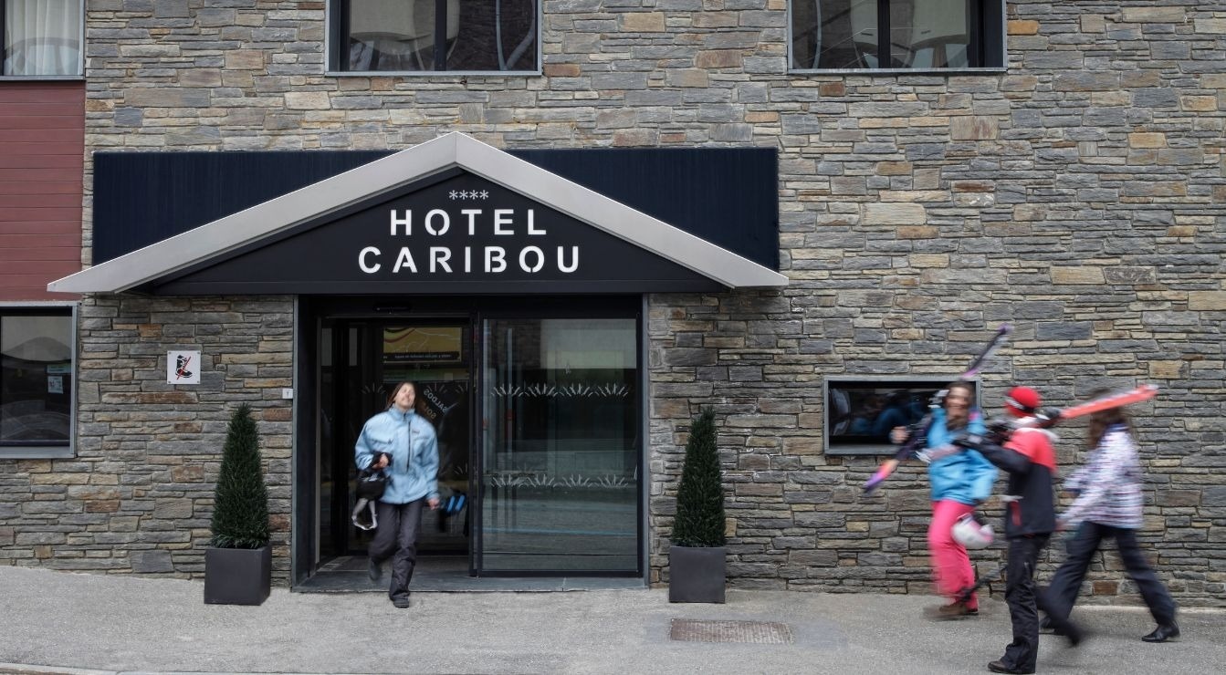 Das Bild zeigt den Eingang des Hotels Caribou, aus dem eine Person heraustritt, während rechts eine Gruppe von Skifahrern mit Bewegungsunschärfe vorbeigeht.