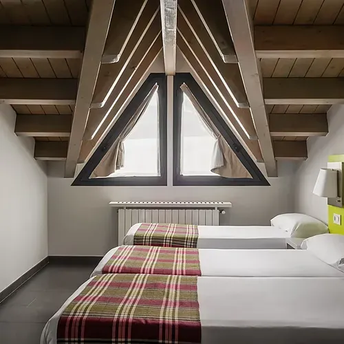 Cozy hotel room with twin beds, vaulted wood ceiling, and large triangular windows. Hotel Cristina | Andorra | Estival Group | Official Web